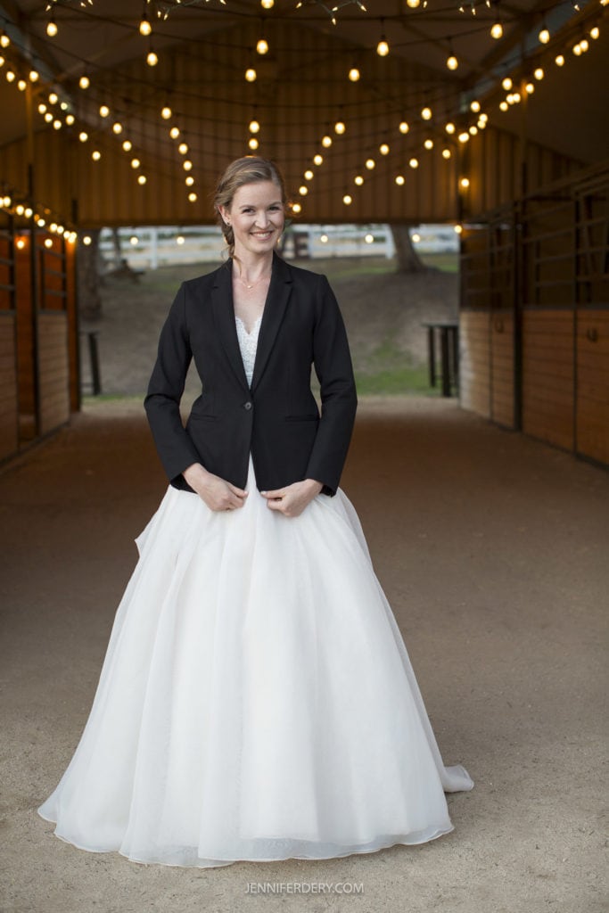 A woman in a white wedding dress and black blazer stands in a barn aisle adorned with string lights. She smiles and holds the edges of her open blazer. The rustic wedding setting includes wooden stable doors and an outdoor view. The image is credited to jenniferdery.com.