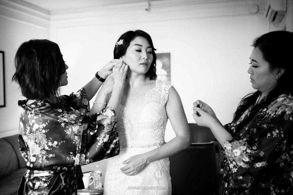 A bride with short, wavy hair adjusts one earring while two women in floral robes assist her with jewelry. The bride is wearing a lace wedding dress, and the scene appears to be set in a well-lit room decorated with minimal furnishings at a Japanese Friendship Garden Wedding.