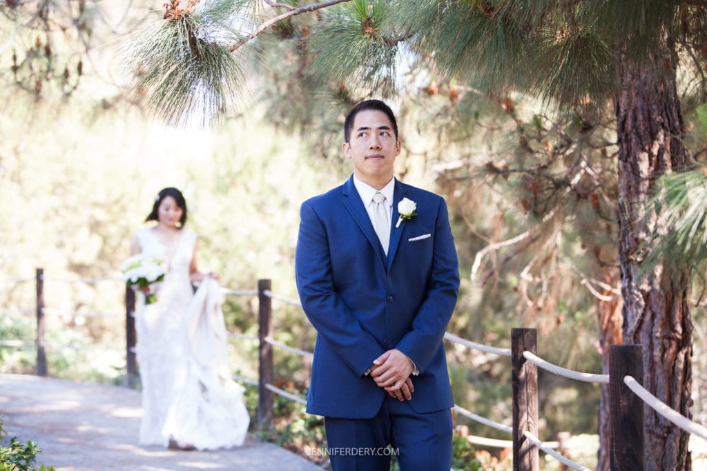 A groom in a blue suit and white boutonniere stands under a tree with a thoughtful expression as a bride in a white wedding dress and veil approaches from behind, holding her dress up slightly. They are outdoors on a pathway surrounded by greenery at the serene Japanese Friendship Garden Wedding.