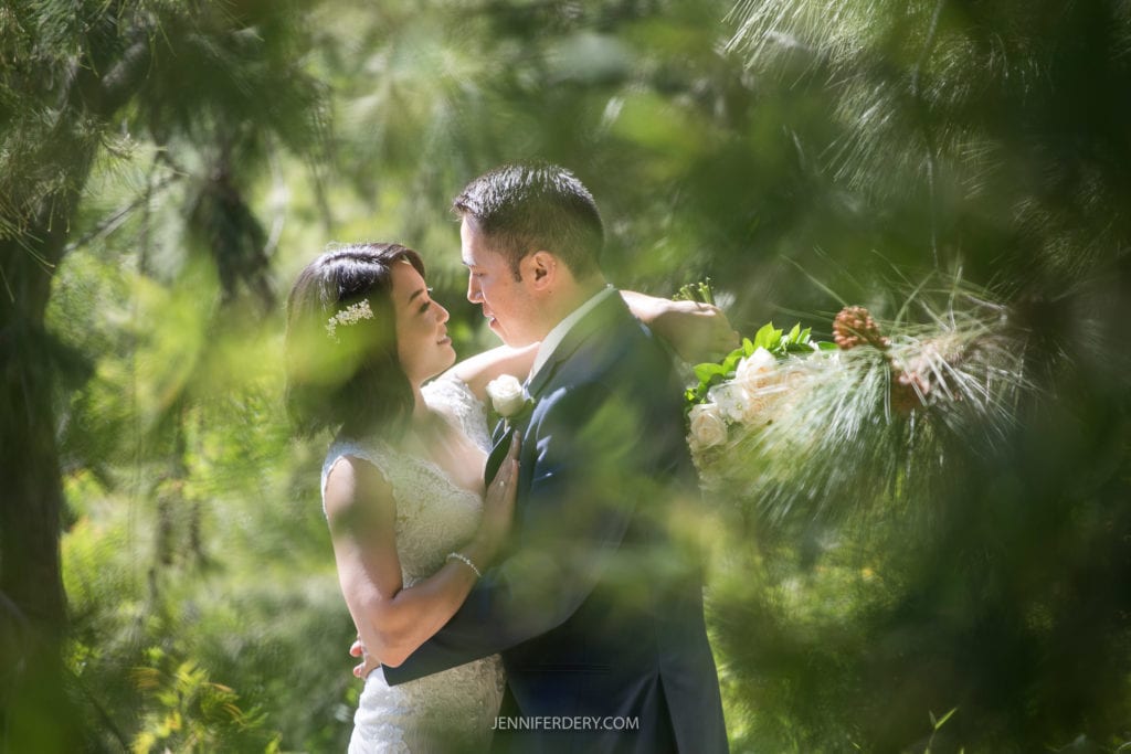 A bride and groom embrace lovingly amidst lush greenery at their Japanese Friendship Garden wedding. The bride holds a bouquet of white roses, and both gaze into each other's eyes as light filters through the trees, creating a serene and romantic atmosphere.