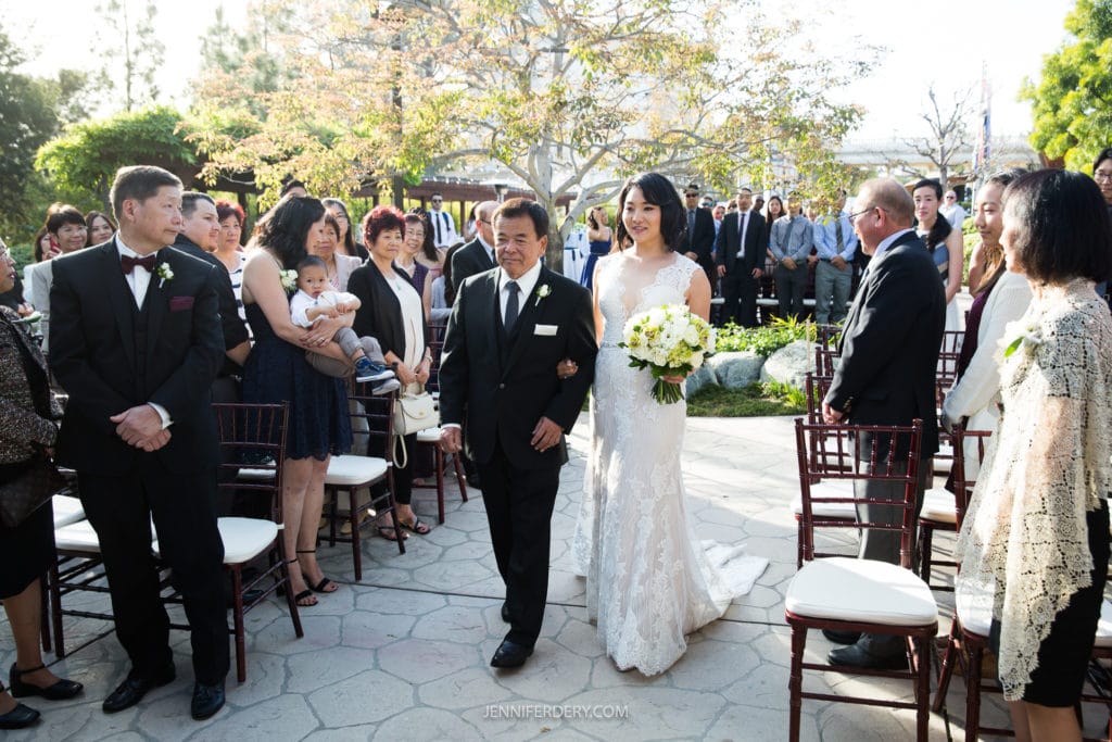 A bride in a white lace gown, holding a bouquet, walks down an outdoor aisle at the Japanese Friendship Garden Wedding, escorted by an older man in a black suit and tie. Guests, some standing and taking photos, watch from either side. Trees and sunlight enhance the serene scene.