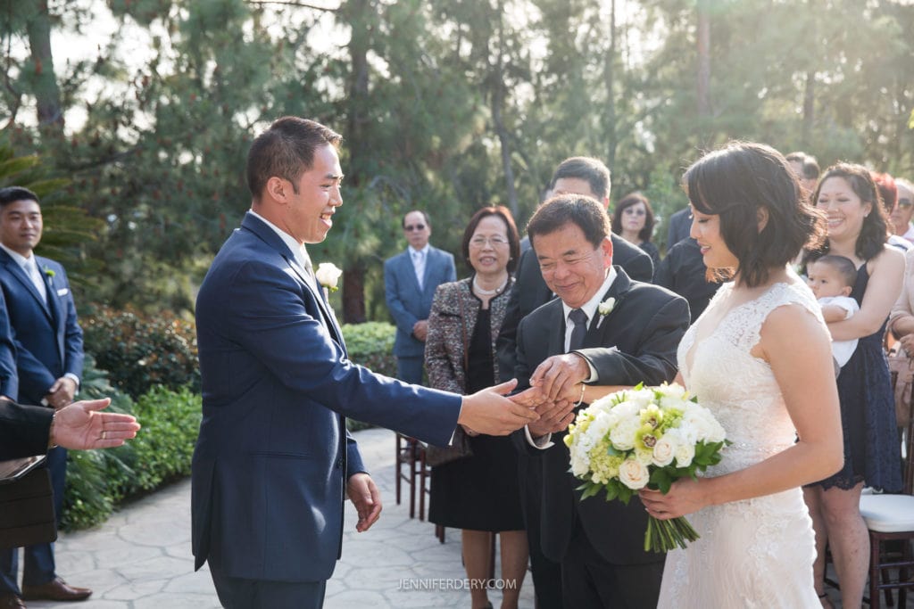 A man in a blue suit shakes hands with another man while a woman in a white wedding dress holding a bouquet looks on, smiling. They are outdoors at the Japanese Friendship Garden, surrounded by trees and guests in formal attire. The setting appears to be a wedding ceremony.