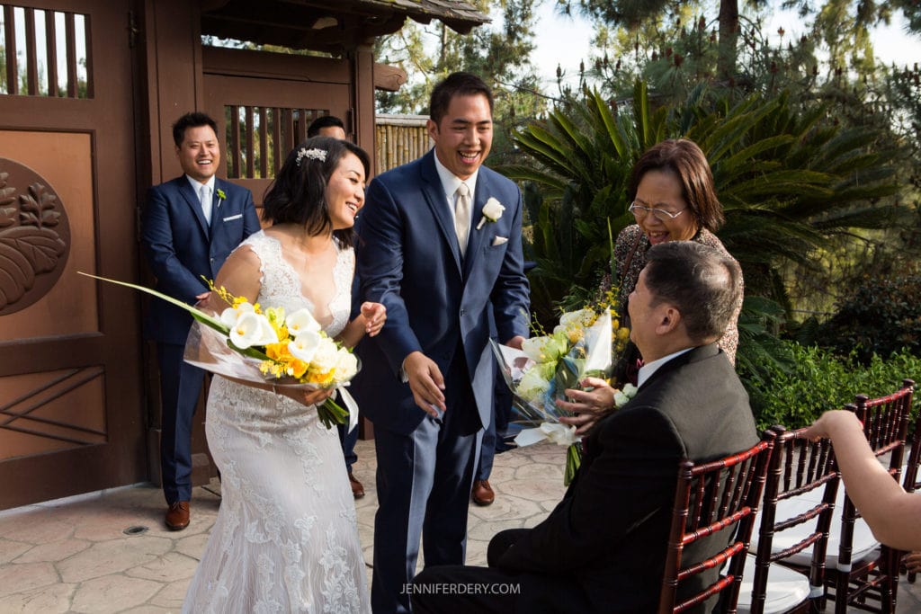 A bride and groom, dressed in a white gown and blue suit respectively, smile and hand flowers to seated guests during an outdoor wedding ceremony at the Japanese Friendship Garden. Two guests, one holding a bouquet, sit in wooden chairs, while two men in suits stand in the background.