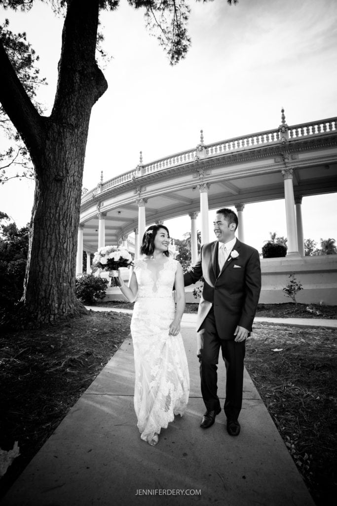A bride and groom walk hand in hand down a path, smiling at each other. The bride, in a lace gown and carrying a bouquet, and the groom, in a suit, seem joyful. The background features tall trees and a grand, columned structure reminiscent of a Japanese Friendship Garden. The image is in black and white.