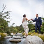 bride and groom crossing a stream using large stepping stones with the scenery of the japanese friendship garden of balboa park as the backdrop
