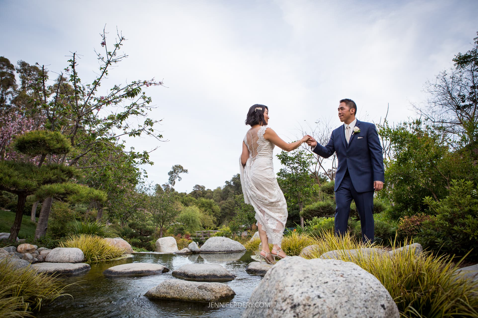 bride and groom crossing a stream using large stepping stones with the scenery of the japanese garden of balboa park as the backdrop