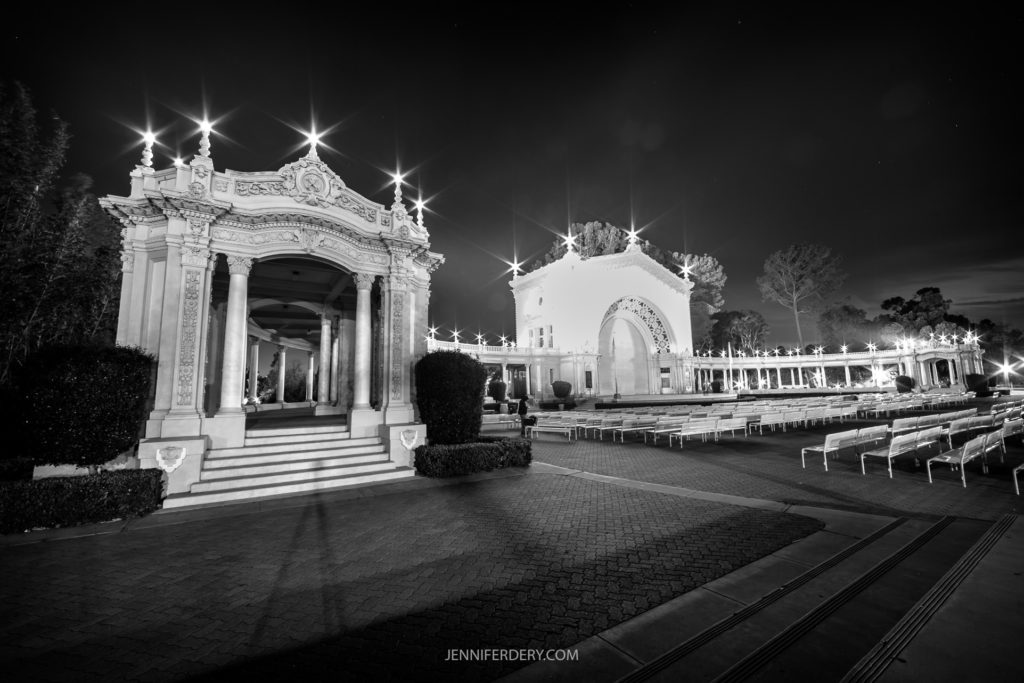 A black and white nighttime image captures an ornate, classical-style building with decorative arches and columns. Stairs lead up to the entrance, flanked by a seating area with rows of benches. In the background, trees and other buildings evoke a serene ambiance at the Organ Pavilion in Balboa Park, CA