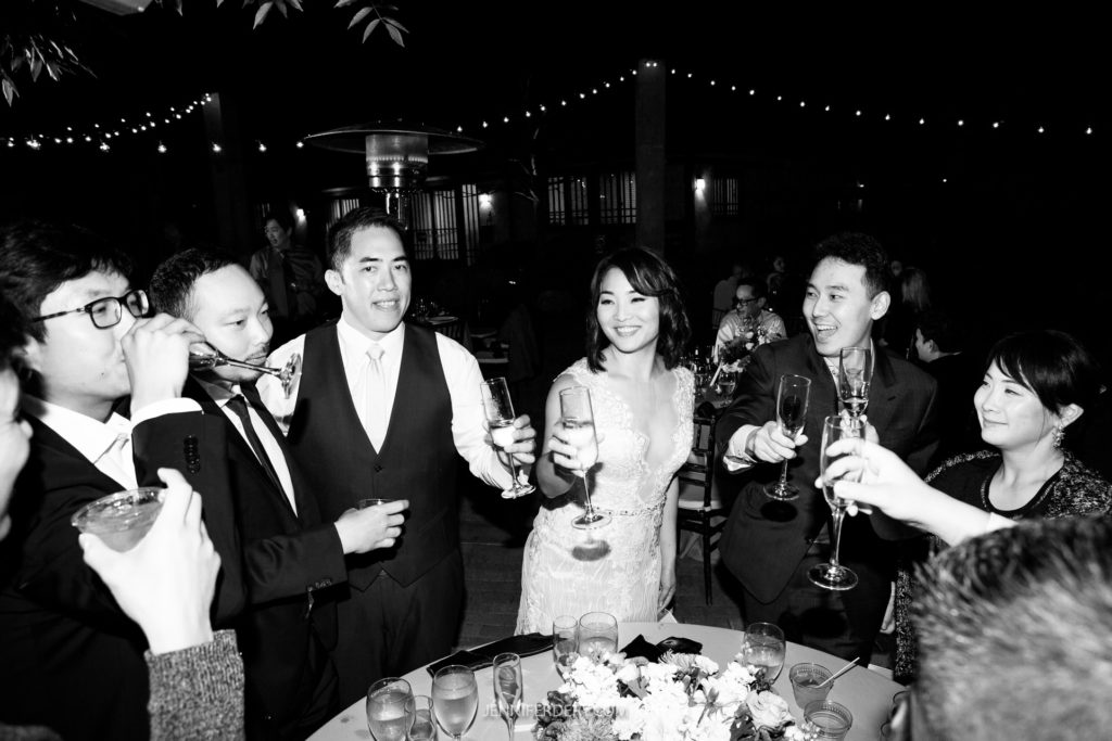 A group of people dressed elegantly are raising their glasses in a celebratory toast. At the center, a bride in a white gown and a groom in a dark suit smile joyfully. The setting, at the Japanese Friendship Garden wedding, is a nighttime event accented with string lights in the background.