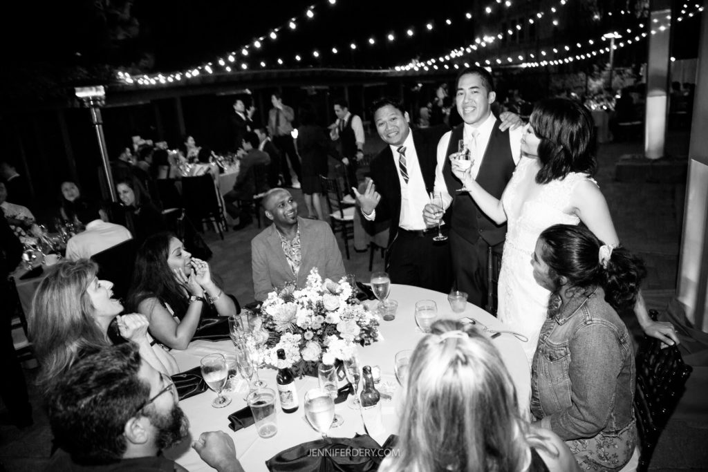 A black and white photo captures a joyous Japanese Friendship Garden wedding reception. Guests are seated around a table, laughing and chatting. The bride and groom stand nearby, smiling and holding drinks, while string lights twinkle in the background, creating a festive atmosphere.