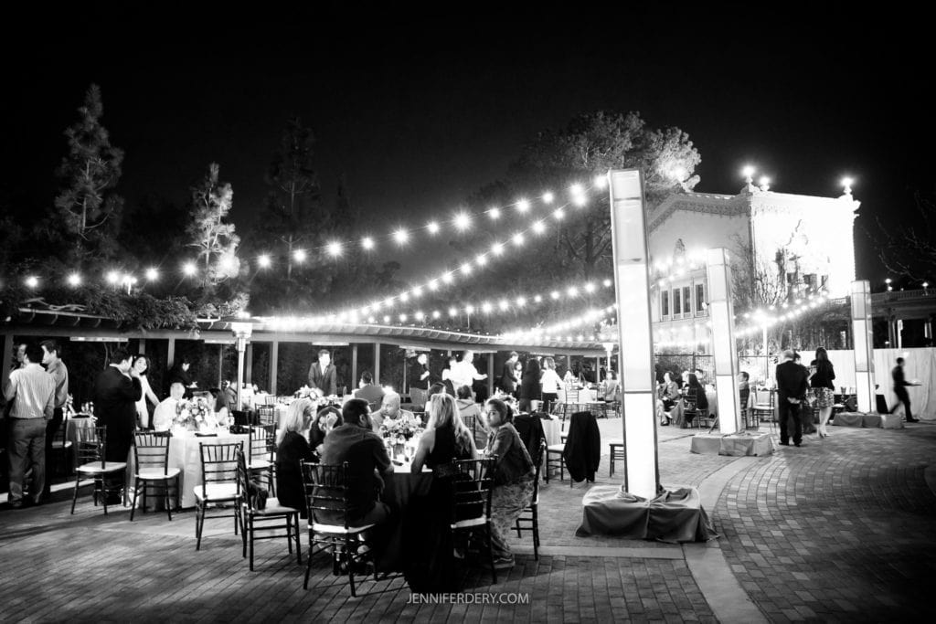 A nighttime outdoor event at the Japanese Friendship Garden Wedding features guests seated at round tables under strings of lights. Some attendees are standing and mingling, while a lit building in the background enhances the ambiance. The photo is in black and white.
