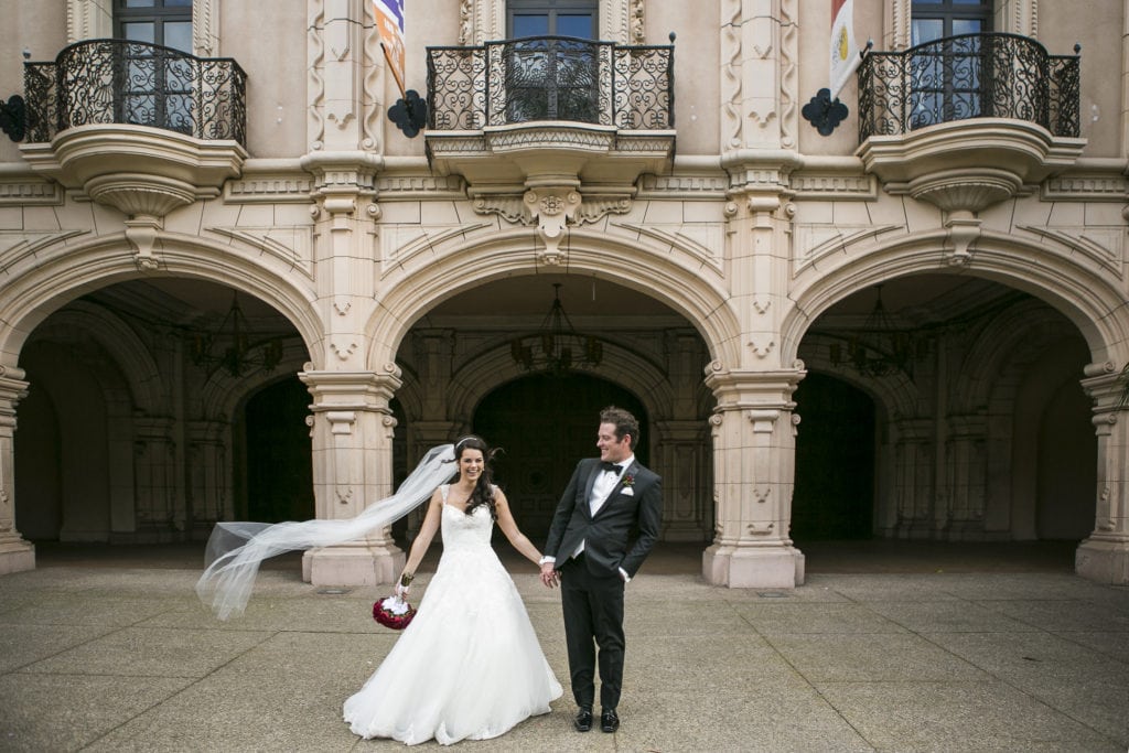 candid wedding couple in front of arches in balboa park. wearing black tie wedding attire and a wind-swept white veil blowing in the wind