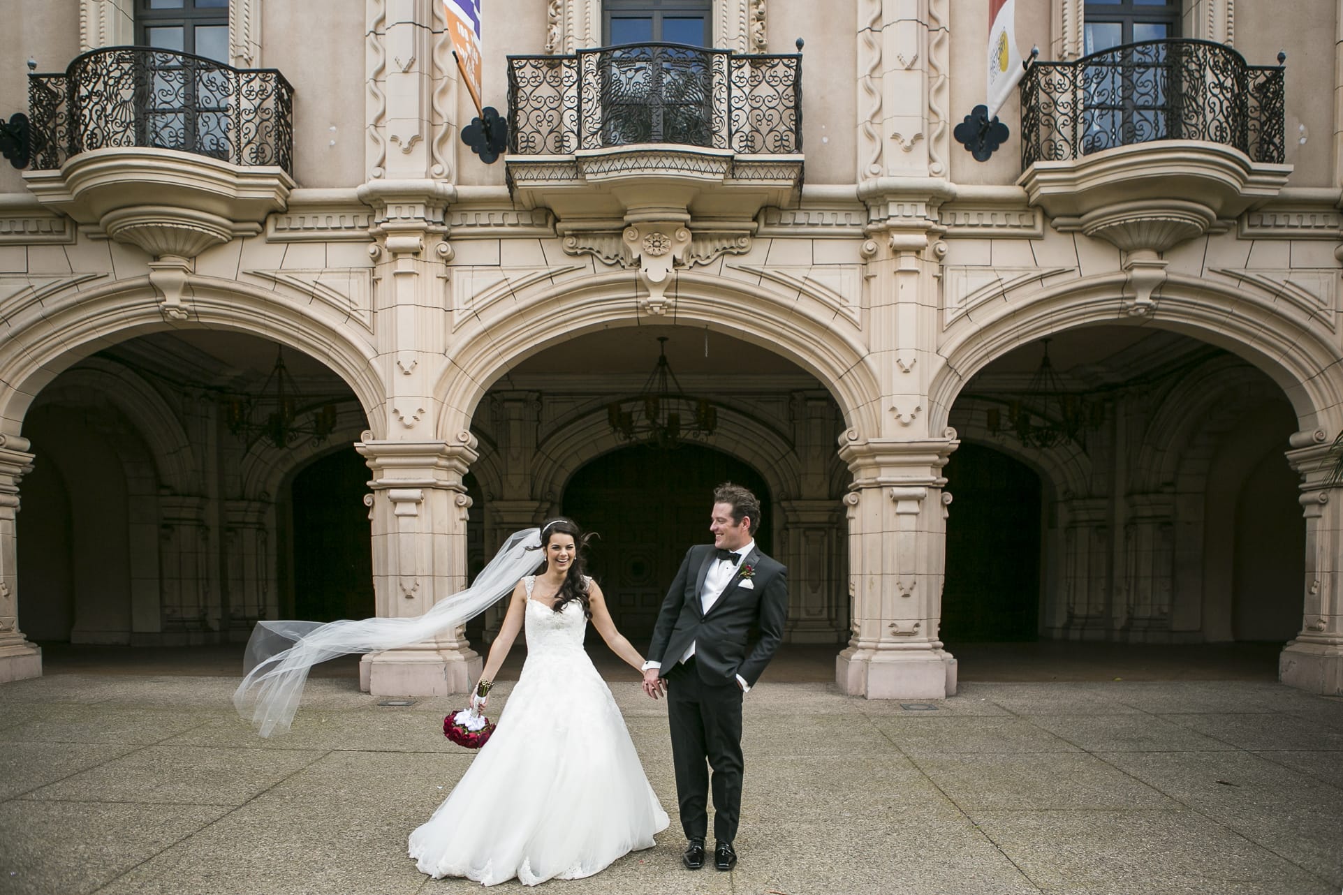 candid photo of a wedding couple in balboa park in black tie wedding attire and a wind-swept white veil blowing in the wind. the brie's vision for photography that she communicated was "silly and fun - yet elegant"