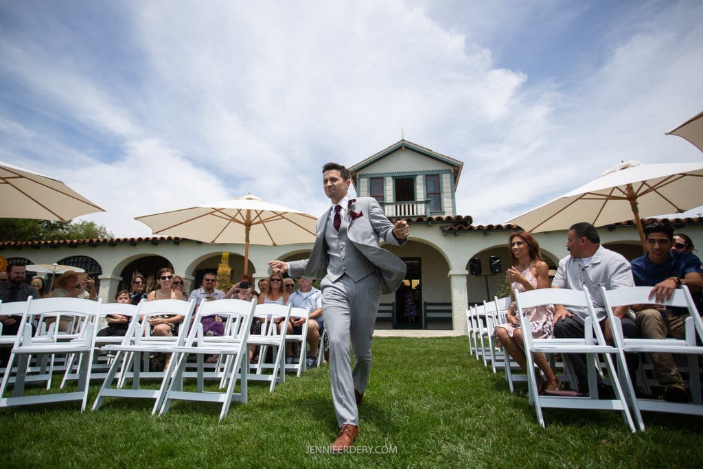 A man in a gray suit walks down an aisle between rows of white chairs with seated guests. The event is outside on a sunny day with a blue sky and a large house in the background. White umbrellas provide shade for some of the guests.