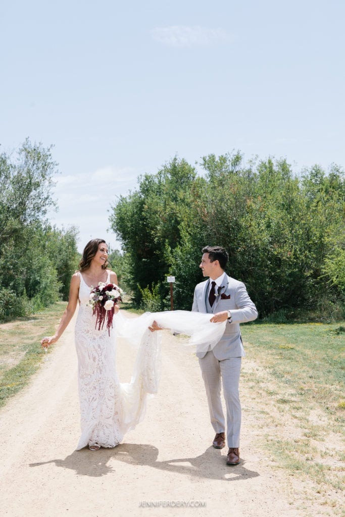 at Rancho Guajome Adobe A bride in a white lace gown and groom in a light gray suit walk down a dirt path surrounded by greenery. The bride holds a bouquet of red and white flowers, smiling, while the groom holds part of her dress, looking at her. The sky is clear and blue.