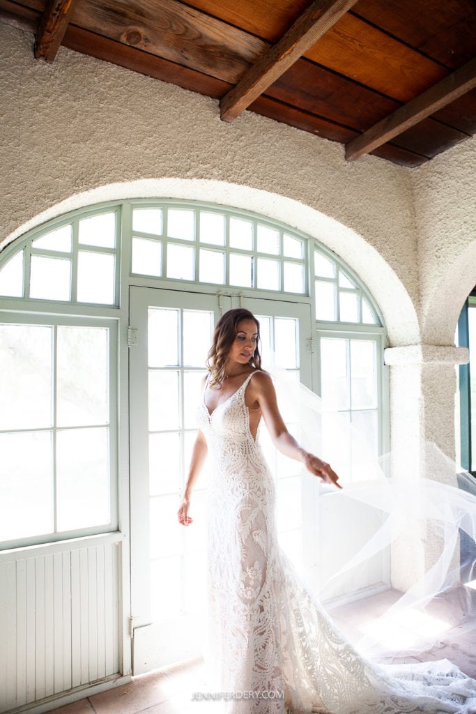 A bride in a white lace wedding dress stands gracefully in front of a set of French doors, illuminated by natural light. She holds her sheer veil with one hand, and her long, wavy hair cascades over her shoulders as she looks to the side.