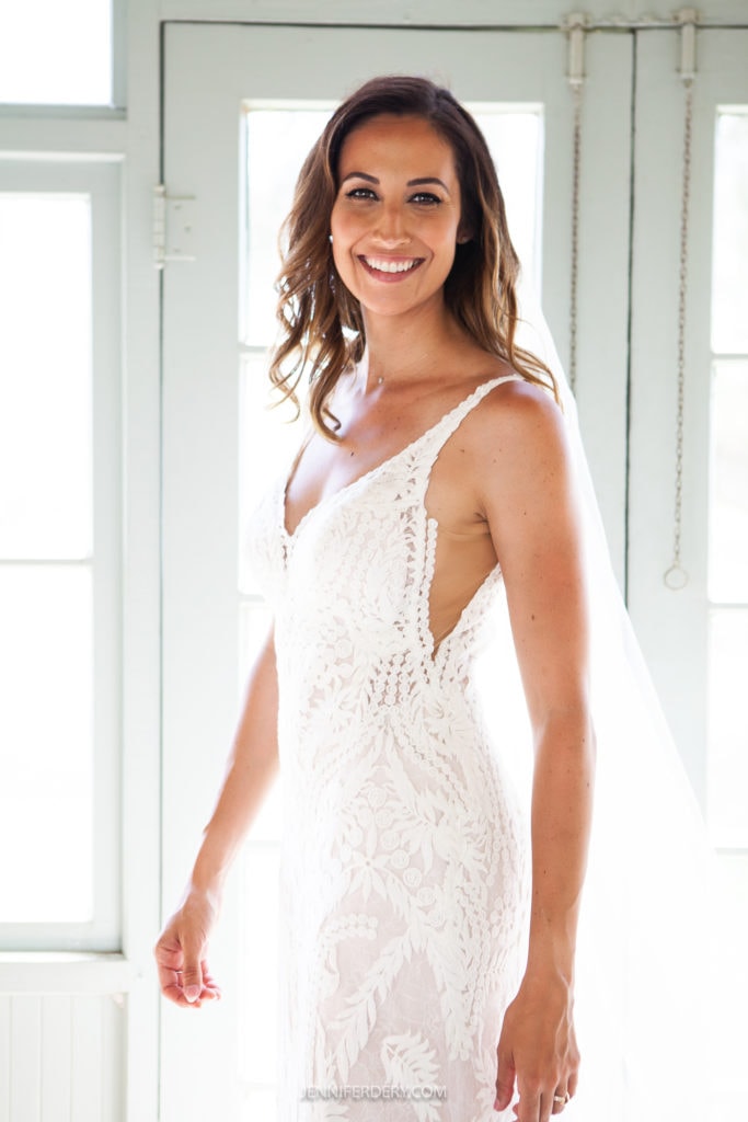 A smiling woman stands indoors, wearing a white lace wedding dress with thin straps and a long veil. She has long, wavy hair and is positioned in front of a door with glass panels, letting in natural light.