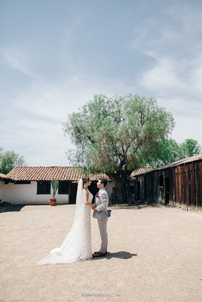 A bride and groom stand outdoors in front of rustic buildings at Rancho Guajome Adobe. The bride, wearing a white dress, and the groom, in a light gray suit, share an intimate moment under a clear sky. A large leafy tree stands in the background.