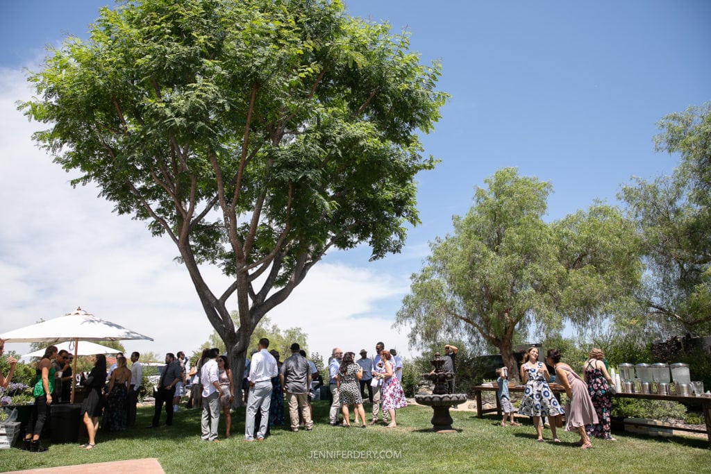 A group of people in formal attire gather outdoors under a large tree at Rancho Guajome Adobe on a sunny day. Tables with refreshments and an umbrella are visible. A clear blue sky and additional trees are in the background. The setting appears to be a garden or park.