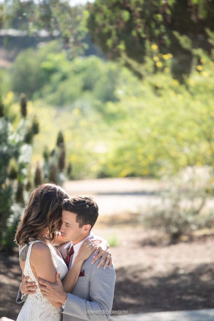 A couple, dressed for their wedding, shares an intimate embrace outdoors at Rancho Guajome Adobe. The bride is wearing a lace wedding dress, and the groom is in a light grey suit. They are surrounded by greenery with sunlight filtering through the trees, creating a serene and romantic atmosphere.