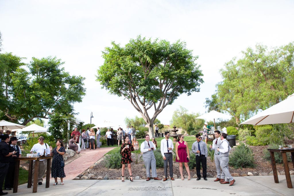 at a wedding reception at Rancho Guajome Adobe A group of people dressed in formal attire are gathered outdoors. Tables with umbrellas are placed around. The scene is lush with green trees and plants, and the sky is clear. People are chatting and mingling in the background.