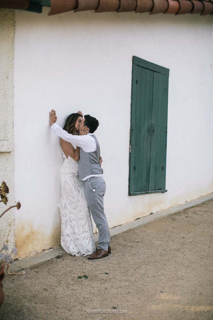 at Rancho Guajome Adobe: A couple shares an intimate moment against a white wall with a green wooden door. The bride is wearing a lacy white wedding dress, and the groom is dressed in a light gray suit and brown shoes. They are embracing passionately, with the bride's hand resting on the wall.