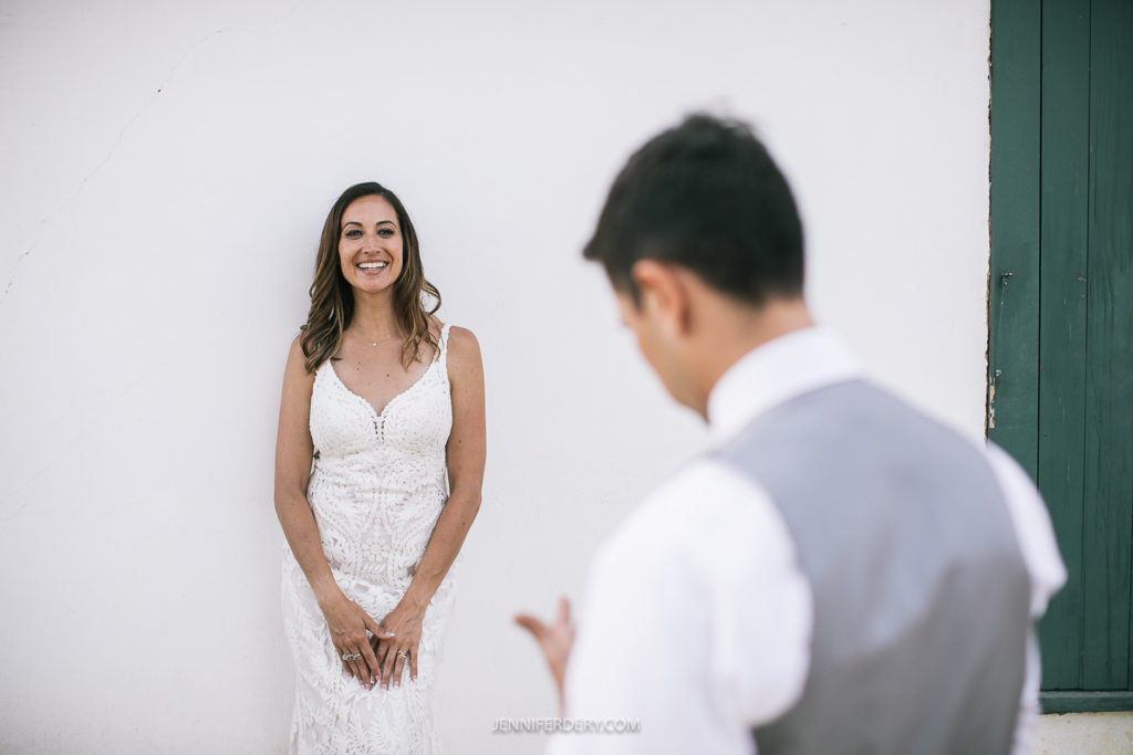 A bride in a white lace dress smiles joyfully against a white wall. In the foreground, a person dressed in a white shirt and gray vest, possibly the groom, is seen from the back. The setting appears bright and airy.