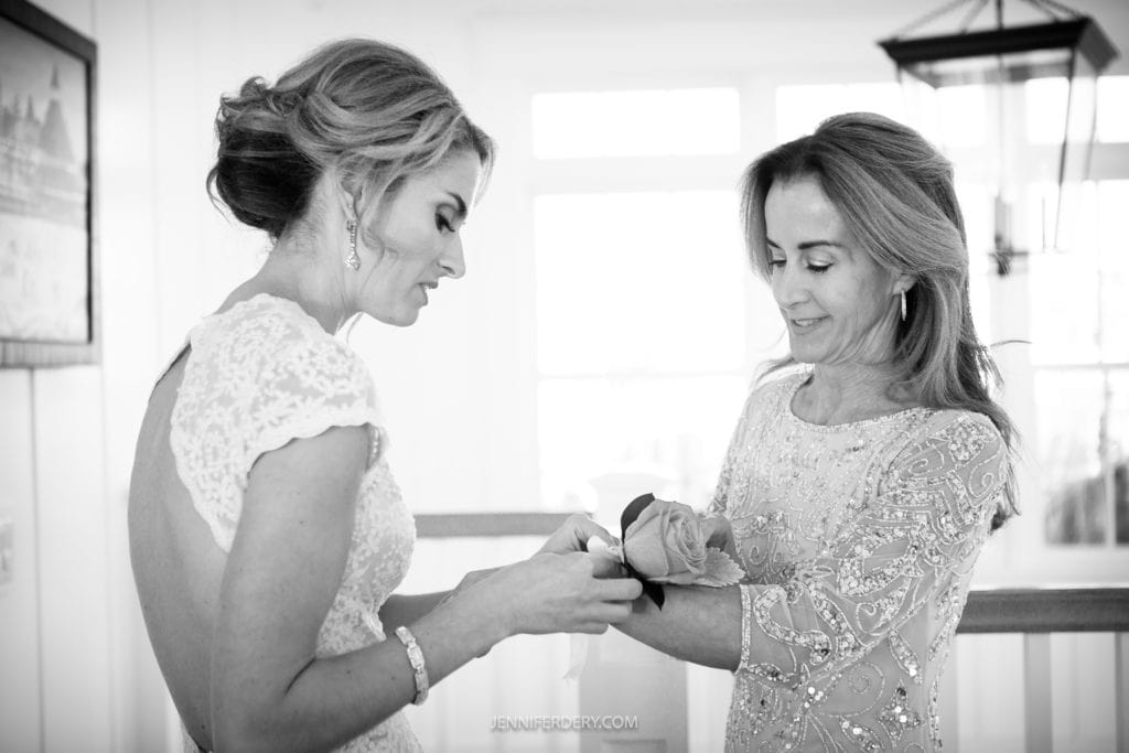 A bride in a lace gown and the elegantly dressed mother of the bride stand indoors. The woman helps the bride adjust something on her wrist, possibly a corsage. They both appear focused on the task, with a window in the background allowing natural light in.