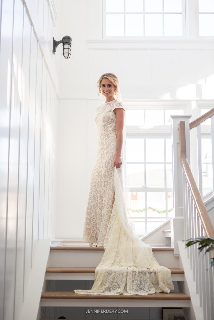 A smiling bride stands on a staircase in a bright room with large windows. She wears a lace wedding gown with a train flowing down the stairs. Sunlight illuminates her from behind, enhancing the details of the gown and her overall radiant appearance.
