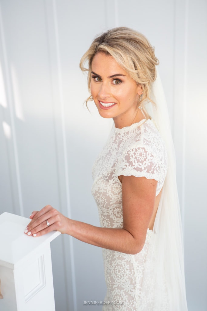 A bride with blonde hair styled in loose waves stands in front of a light-colored background. She is wearing a white lace wedding gown with cap sleeves and a back cutout. A sheer veil falls down her back, and she is smiling while looking over her shoulder.