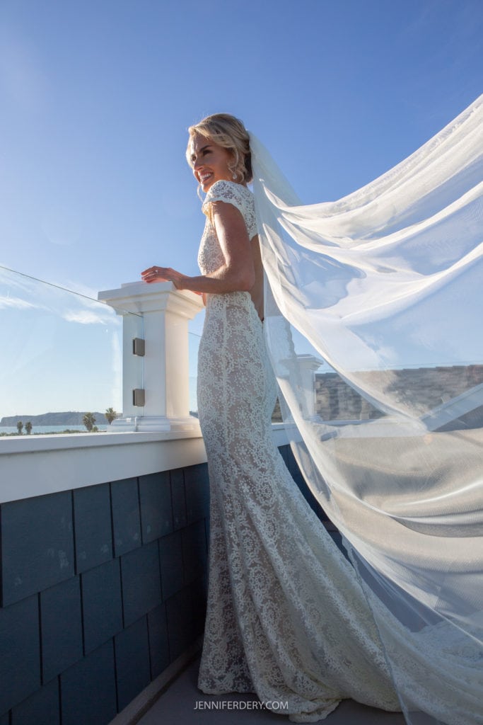 A bride in a lace wedding dress and long flowing veil stands on a rooftop patio, leaning on the glass railing and smiling happily with a clear blue sky in the background.
