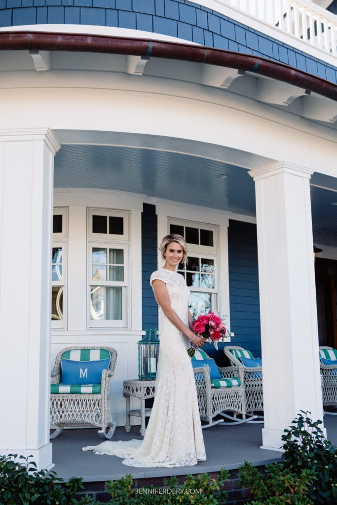 A bride in an elegant lace wedding dress stands on the porch of a house with blue siding. She holds a vibrant bouquet of pink flowers and smiles. The porch has white columns, wicker chairs with cushions, and is decorated in coastal style.