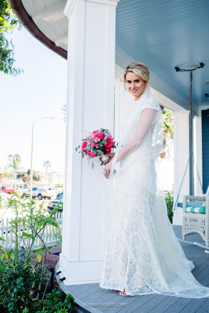 A bride in a white lace wedding dress and veil stands on a porch, holding a vibrant bouquet of pink and white flowers. She smiles softly, surrounded by greenery and a white picket fence. The house is painted blue with a porch swing visible in the background.