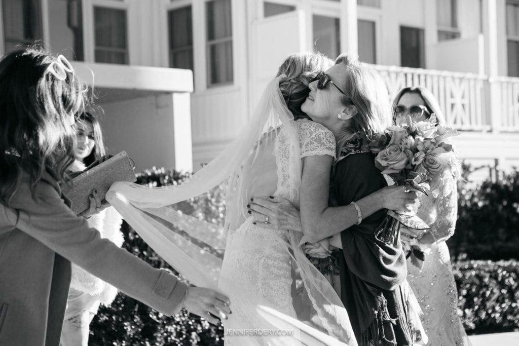 A black and white photo shows a bride with a long veil hugging an older woman, possibly her mother, who is holding a bouquet of flowers. Two other women are visible nearby, one holding a notebook or planner. The background features a building with large windows.