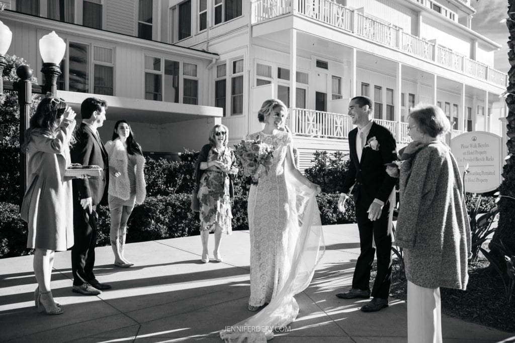 A bride in a long, flowing white dress with a veil and a bouquet stands next to the groom in a suit. They are outdoors, surrounded by five people, with a large, elegant building in the background. Everyone appears to be smiling and interacting in a joyful manner.