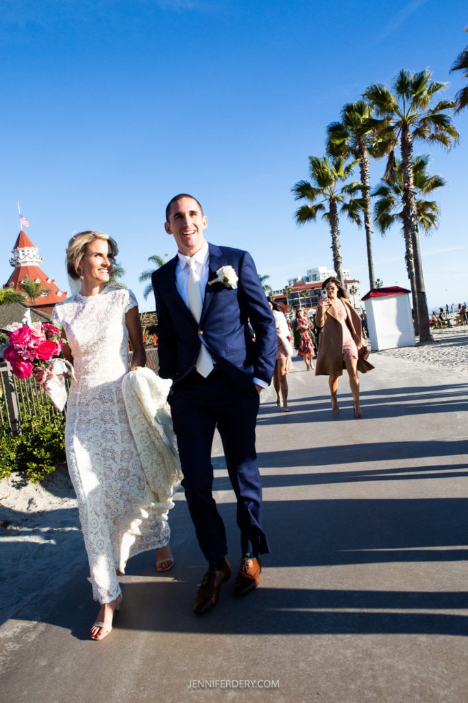 A bride and groom walk on a sunny boardwalk by the beach. The bride wears a lacy white dress and carries pink flowers, while the groom is in a navy suit with a white boutonniere. Palm trees line the path, and a woman in a tan dress follows them.