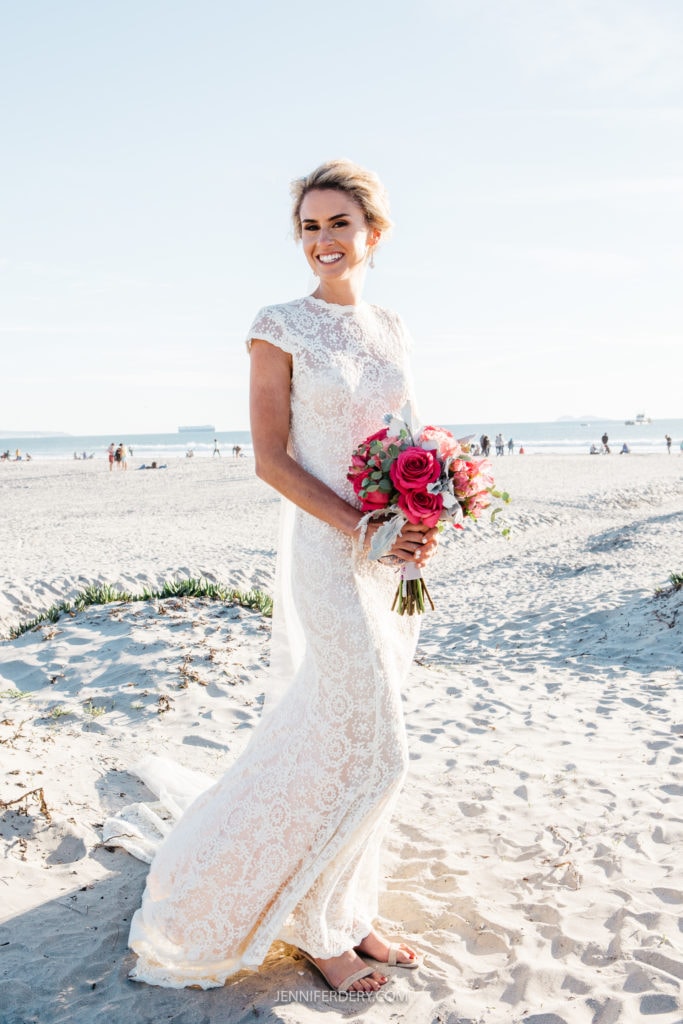 A bride stands on a sandy beach, smiling while holding a bouquet of pink, red, and white flowers. She wears a white lace wedding dress with short sleeves and a train. The background shows the ocean, ships, and people walking on the beach.