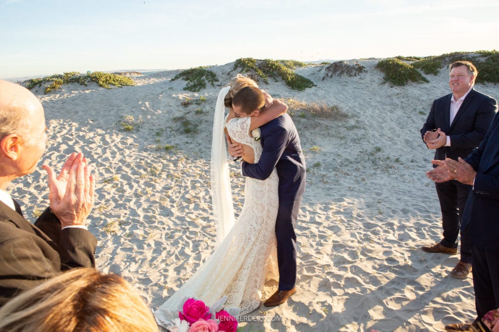 A bride and groom share an embrace during their wedding ceremony on a sandy beach, surrounded by a few onlookers. The bride wears a white lace gown and a veil, and the groom is in a navy suit. The setting is bright and sunny, with dunes and greenery in the background.