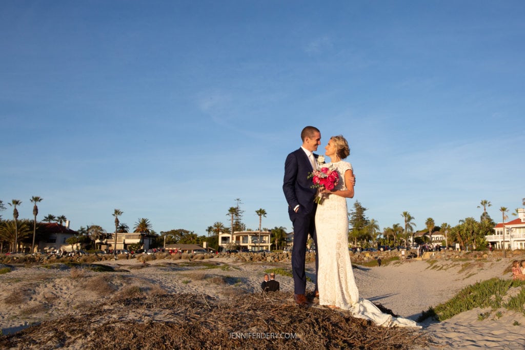 A bride and groom stand on a sandy dune at a beach, dressed in formal wedding attire. The bride holds a colorful bouquet, and both are gazing at each other. Behind them, the clear blue sky, scattered palm trees, and distant buildings enhance the serene backdrop.
