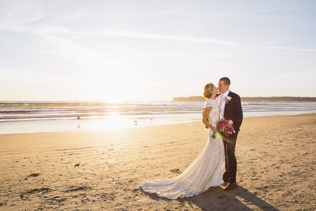 A bride and groom share a kiss on a sandy beach at sunset. The bride wears a long, lace wedding gown and holds a bouquet of flowers, while the groom wears a suit and tie. The sun sets over the ocean in the background, casting a warm glow over the scene.