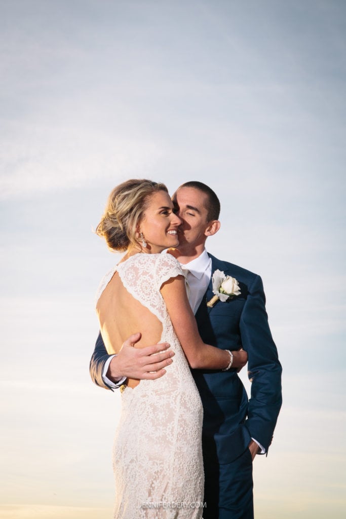 A couple stands close together, smiling and embracing under a clear sky. The woman wears a white lace gown, and the man is in a dark suit with a white boutonniere. They gaze lovingly at each other, highlighting the joyful moment.