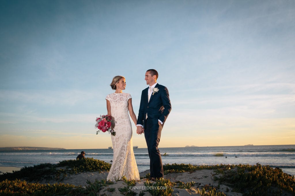 A couple stands hand-in-hand on a beach at sunset. The woman wears a white lacy wedding dress and holds a bouquet of pink flowers. The man is dressed in a blue suit with a white boutonniere. The sky is clear with a warm, golden glow. Waves gently lap in the background.