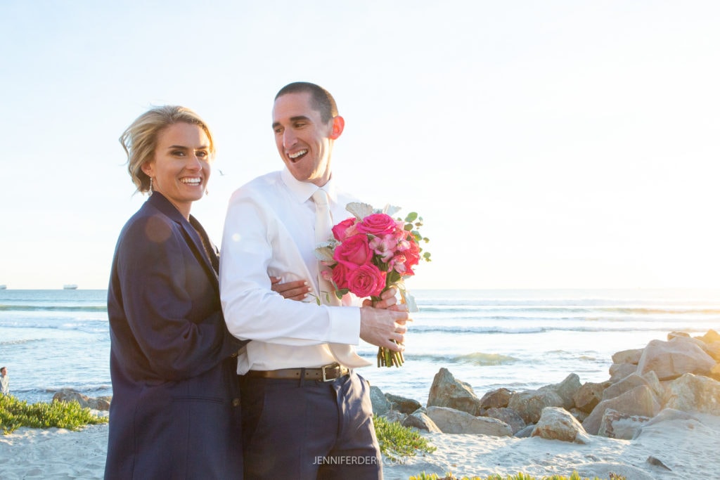 A joyful couple stands by the seaside; the woman wearing a dark blazer hugs the man from behind, who is holding a bouquet of pink and white flowers. The sun is setting, casting a warm glow over the scene with waves and rocks in the background.