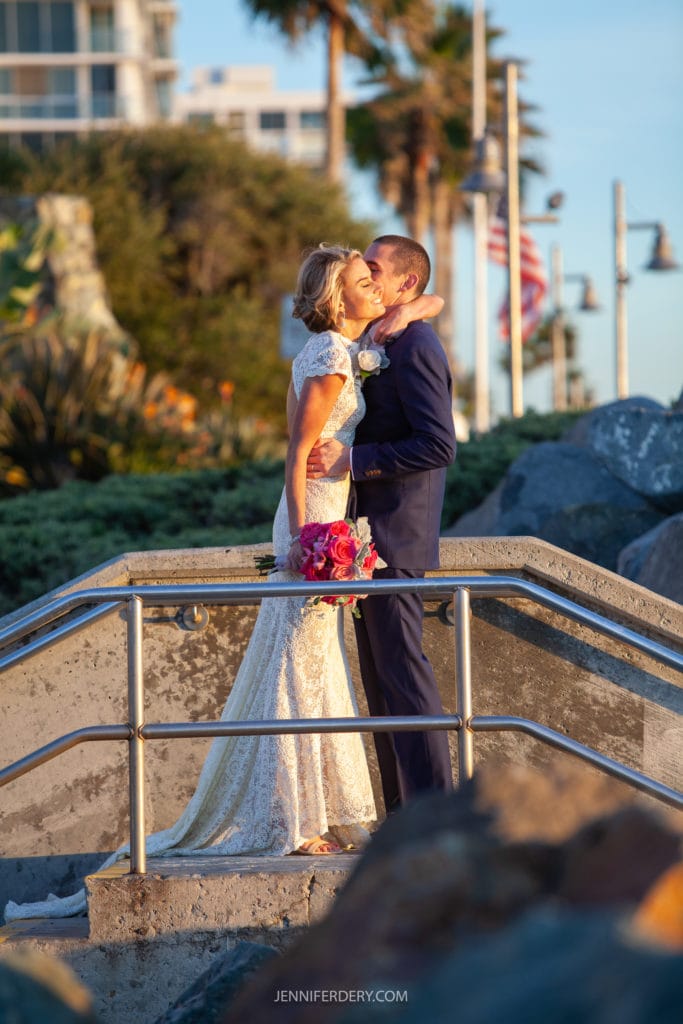 A bride and groom share a kiss on a sunny outdoor walkway. The bride is wearing a white lace gown and holding a bouquet of pink flowers, while the groom is dressed in a navy suit. They are surrounded by greenery, rocks, and American flags fluttering in the background.