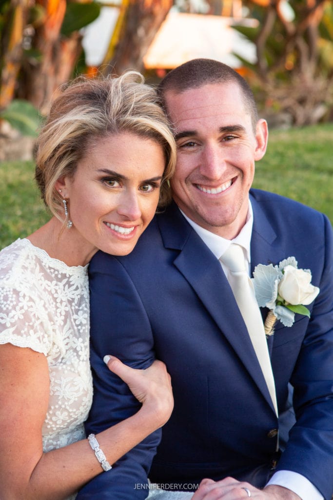 A smiling couple poses closely together outdoors. The woman, wearing a white lace dress, has short blond hair and is leaning on the man, who is wearing a navy blue suit with a white shirt and tie, and a boutonnière on his lapel. Trees and grass are in the background.