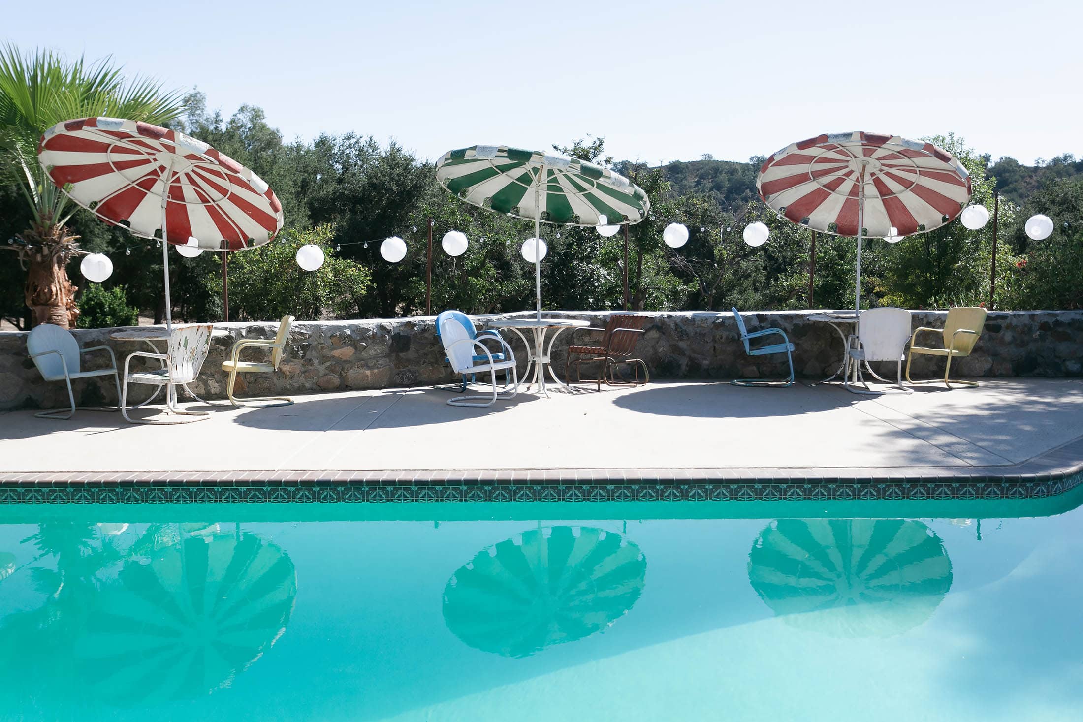 A serene outdoor pool scene, perfect for an intimate elopement, with a row of empty chairs and tables under colorful striped umbrellas. The pool reflects the umbrellas, and string lights add charm to the stone wall backdrop lined with greenery.