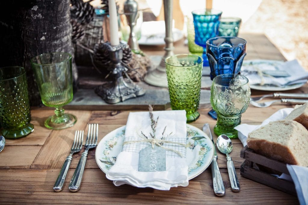 A rustic table setting ideal for an intimate elopement reception features vintage plates, green and blue glassware, silver cutlery, and a bread basket. A white napkin with a handwritten name tag and a sprig of herbs rests on each wooden plate.