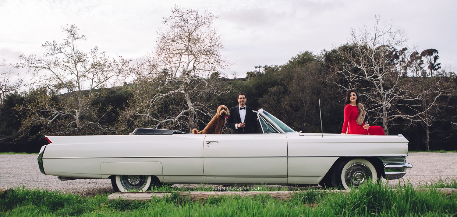 engagement session ideas san diego example of a couple getting engagement photos done with a white classic car in a hollywood glamour style with red dress and black tux. photo includes a dog.