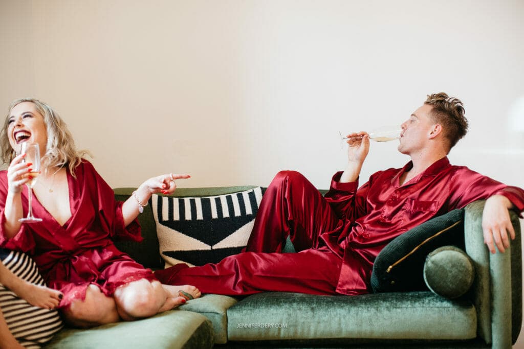 bridal party laying on a green couch in red silk pajama and drinking champagne