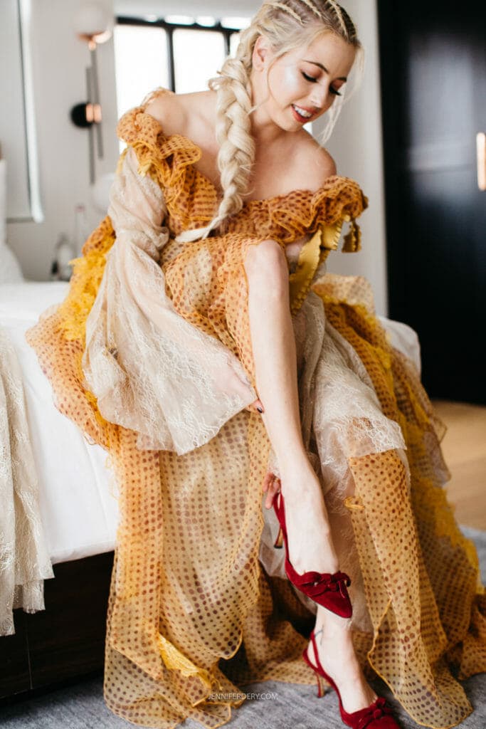 A bride with long, braided blonde hair sits on a bed, gracefully adjusting one of her red high heels. She is wearing an elegant, off-the-shoulder, yellow and beige textured gown and smiling softly. The room is modern and well-lit, with black and white decor.