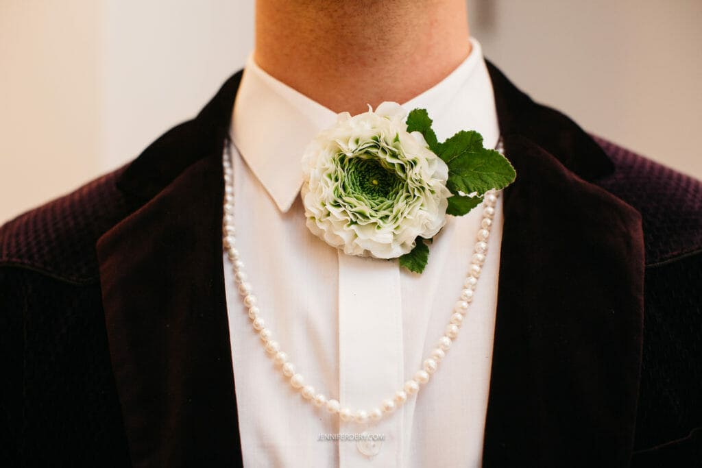 A person wearing a white dress shirt and dark blazer is adorned with a white flower boutonniere pinned to the shirt's collar. The individual also wears a string of pearls around their neck. The background is blurred.